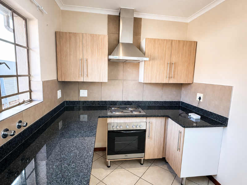 A clean, modern kitchen featuring granite countertops, light wood-finish cabinetry, a built-in oven with electric hob, and a stainless steel extractor fan.