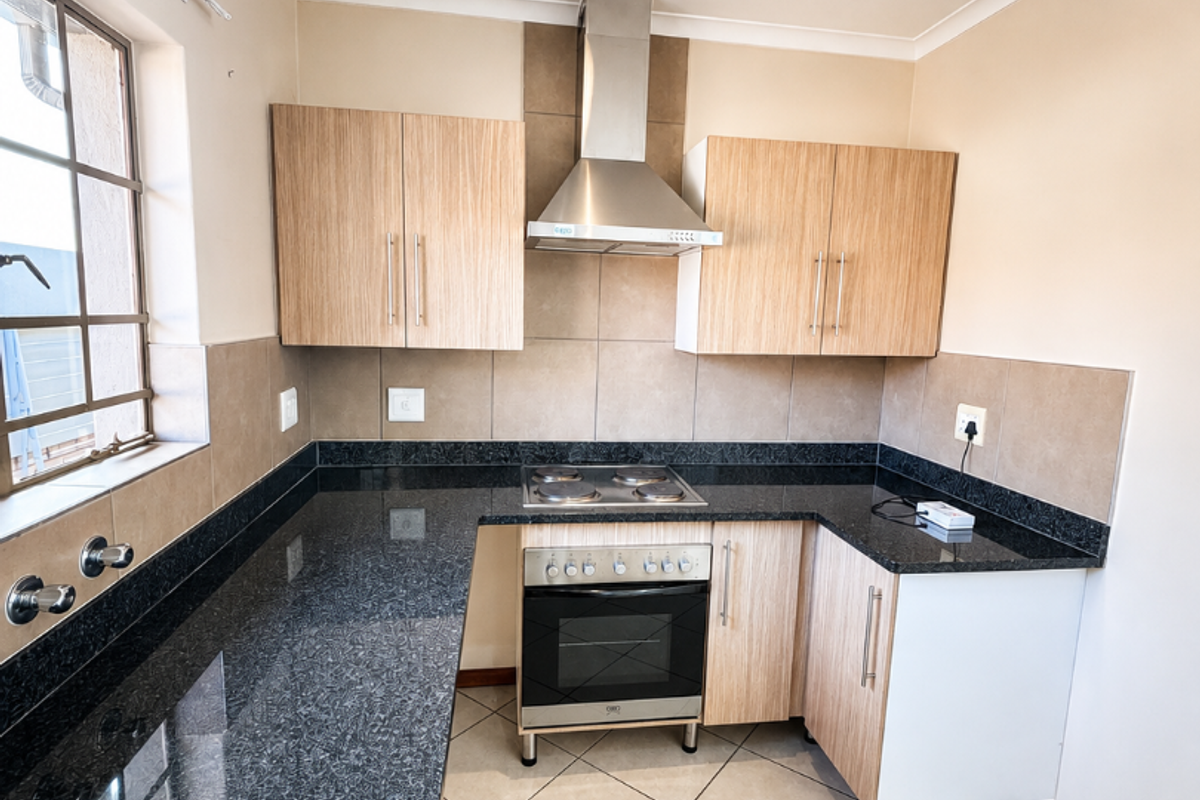 A clean, modern kitchen featuring granite countertops, light wood-finish cabinetry, a built-in oven with electric hob, and a stainless steel extractor fan.
