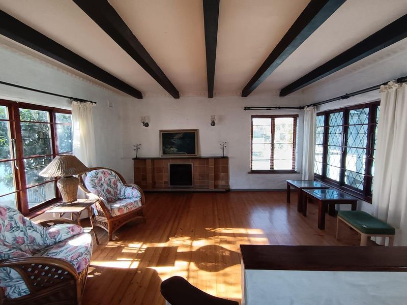 Spacious living room featuring exposed timber ceiling beams, polished timber flooring, a tiled fireplace, and large leaded glass windows providing ample natural light.