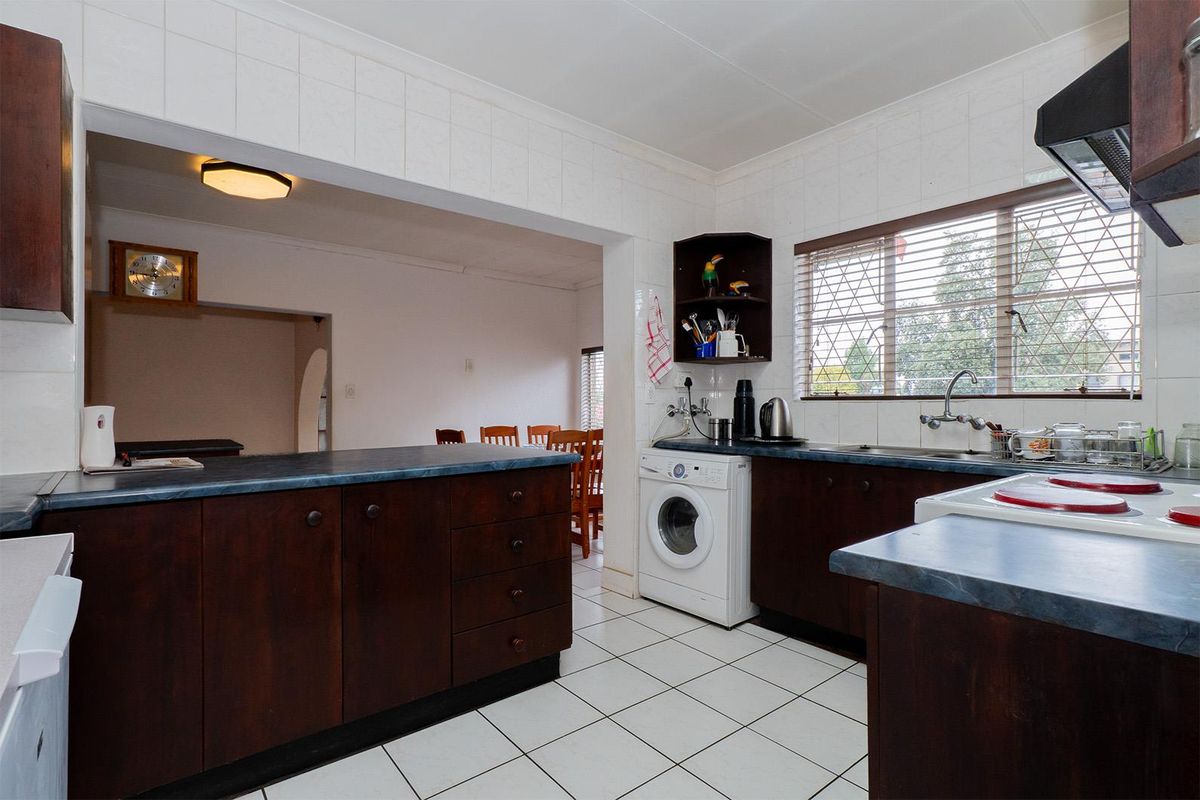 A functional kitchen featuring dark wood cabinetry, laminate countertops, tiled flooring, and an integrated laundry appliance space, opening into an adjacent dining area.