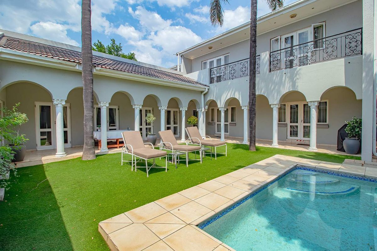 A spacious rear courtyard featuring a swimming pool, artificial lawn, and a covered patio area with classic architectural columns and balcony access.