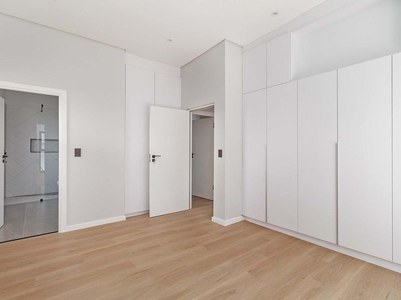 A bright, modern bedroom featuring light wood-look laminate flooring, extensive floor-to-ceiling built-in cupboards, and direct access to an en-suite bathroom.