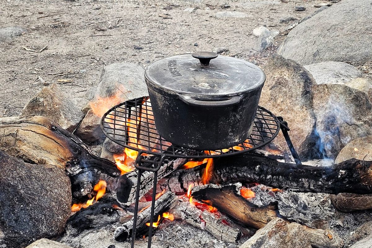 An outdoor campfire setup featuring a metal cooking grate and a cast iron pot, situated on a natural stone and dirt surface.