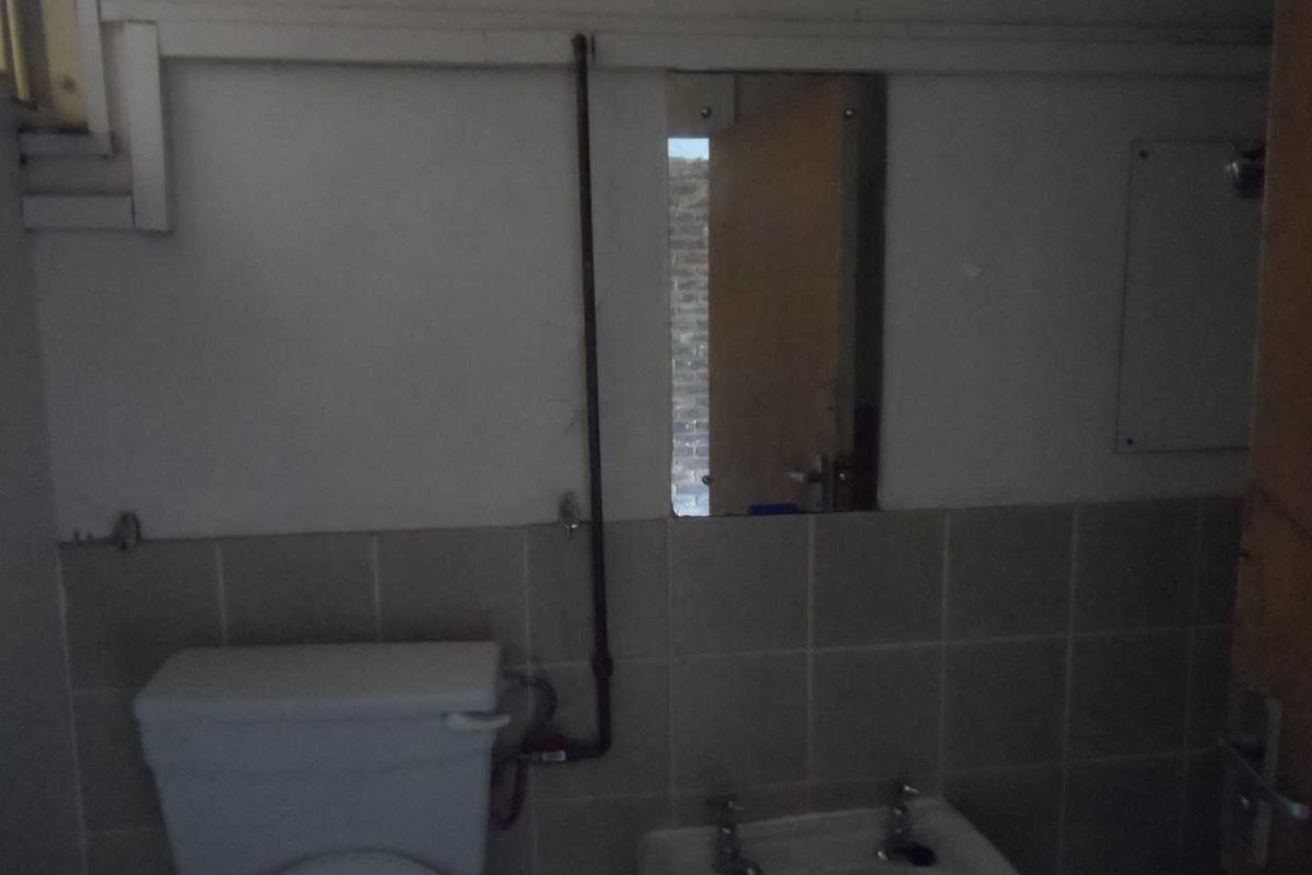 A small, dated bathroom featuring a basic toilet and basin with exposed plumbing and tiled splashback, requiring modernization and improved lighting.