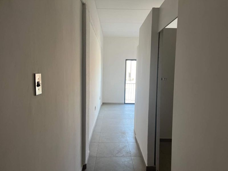 A vacant hallway featuring tiled flooring and plain white walls, leading towards an exterior doorway.