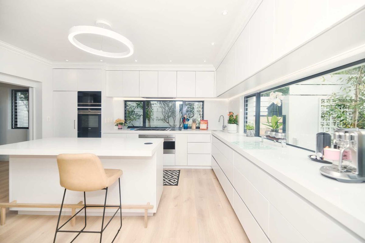 Modern, minimalist kitchen featuring sleek white cabinetry, a central island, integrated appliances, and expansive windows overlooking the garden.