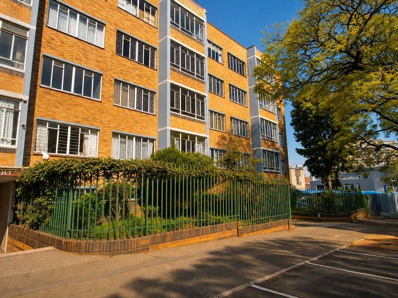 Exterior view of a multi-story brick apartment building featuring metal security fencing and established landscaping along the front facade.