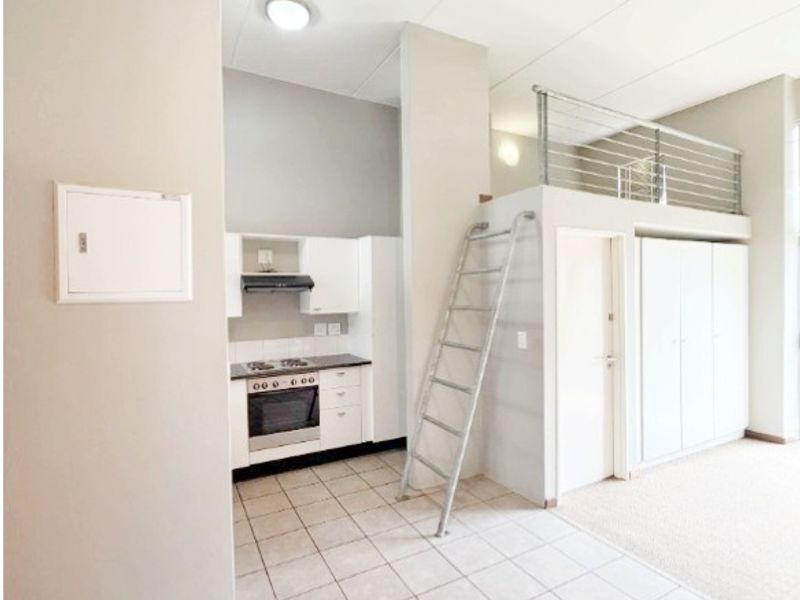Open-plan apartment interior featuring a compact kitchen with built-in appliances, tiled flooring, and a mezzanine level accessible by a fixed metal ladder.
