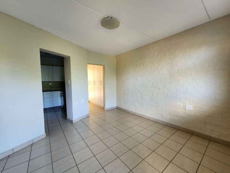 An empty living area featuring tiled floors and a textured brick feature wall, with direct access to the kitchen and bedroom hallway.