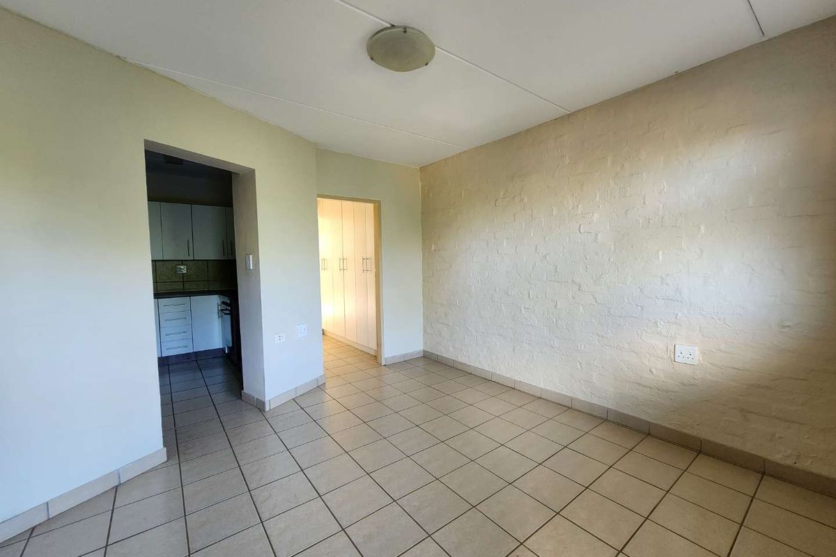 An empty living area featuring tiled floors and a textured brick feature wall, with direct access to the kitchen and bedroom hallway.