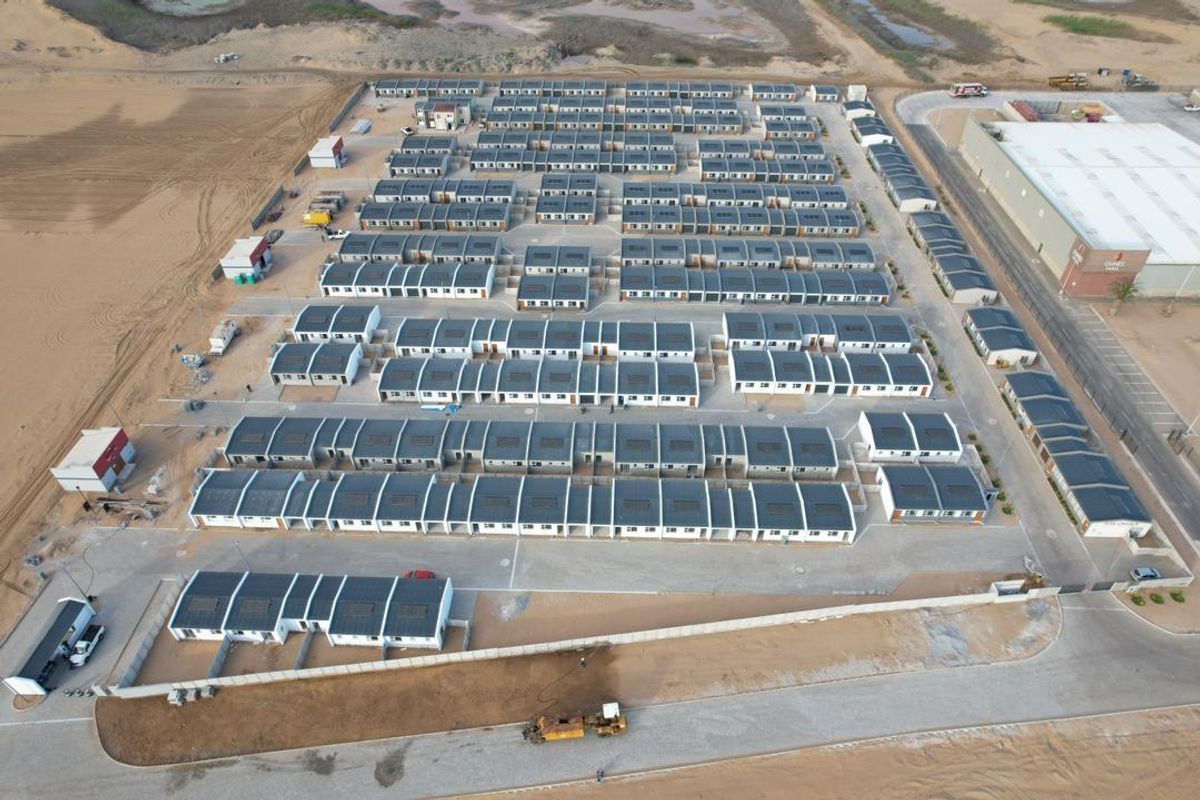Aerial view of a modern townhouse development complex featuring uniform residential units, paved access roads, and perimeter security fencing in a developing area.