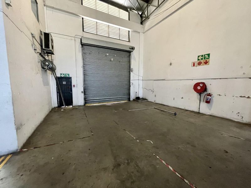 Spacious industrial garage unit featuring a large roller shutter door, concrete flooring, and essential fire safety equipment, suitable for storage or workshop use.