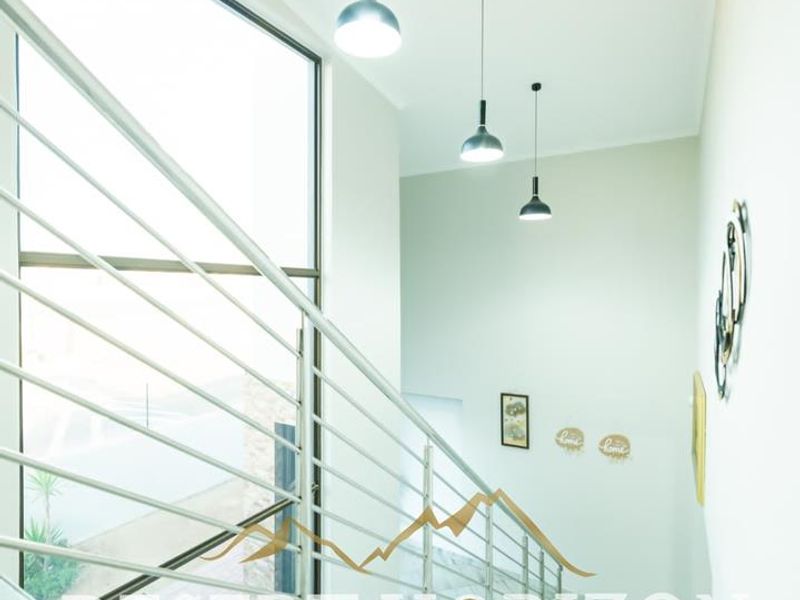 A bright, modern staircase area featuring a sleek metal balustrade, large floor-to-ceiling window, and contemporary pendant lighting.