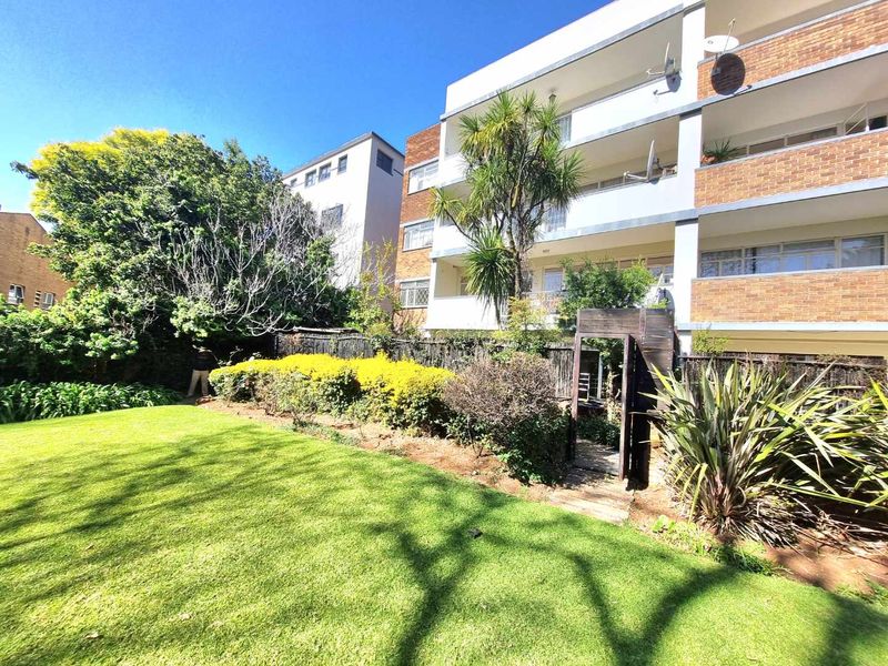A view of the rear apartment building facade featuring balconies, overlooking a well-maintained communal garden area with mature trees and landscaping.