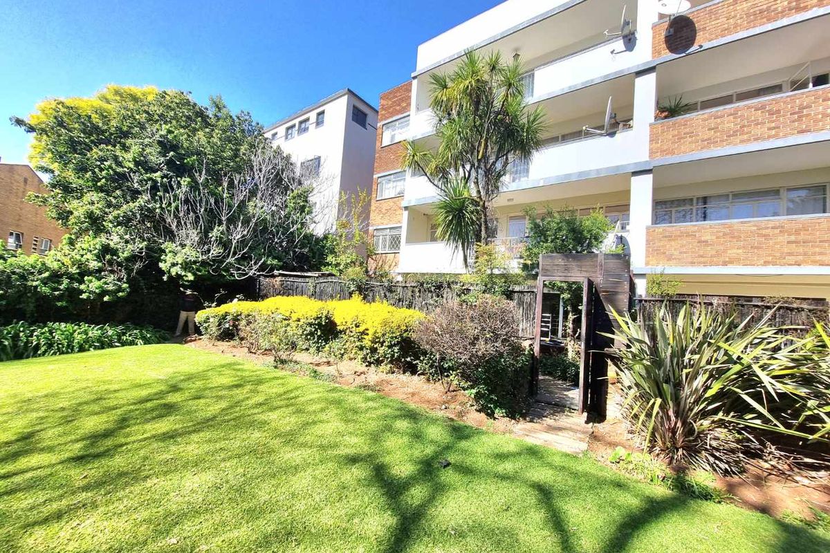 A view of the rear apartment building facade featuring balconies, overlooking a well-maintained communal garden area with mature trees and landscaping.