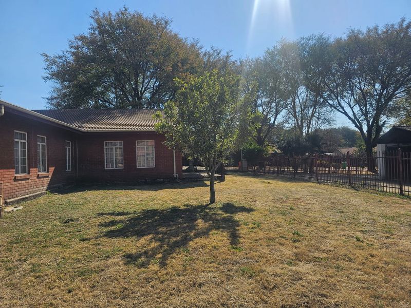 A view of the rear garden featuring a brick house exterior, tiled roof, and metal perimeter fencing.