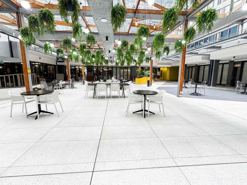 A modern, open-plan commercial atrium featuring a glass roof, exposed timber beams, and high-quality terrazzo flooring, creating a bright and inviting communal space.