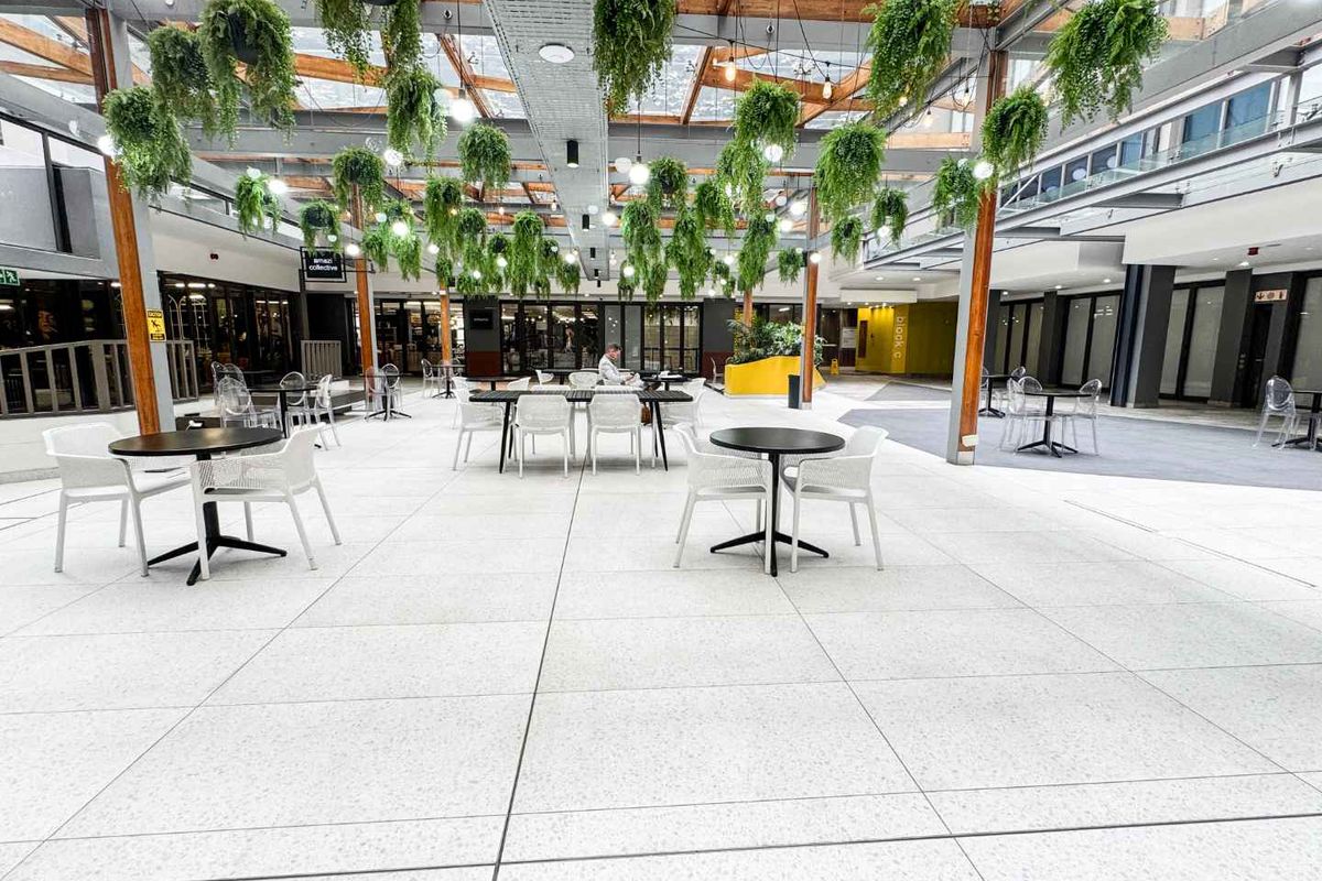 A modern, open-plan commercial atrium featuring a glass roof, exposed timber beams, and high-quality terrazzo flooring, creating a bright and inviting communal space.