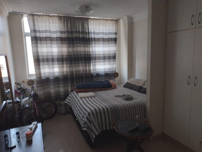 A bedroom featuring built-in cupboards, tiled flooring, and large windows with curtains, offering a functional space with potential for modernization.