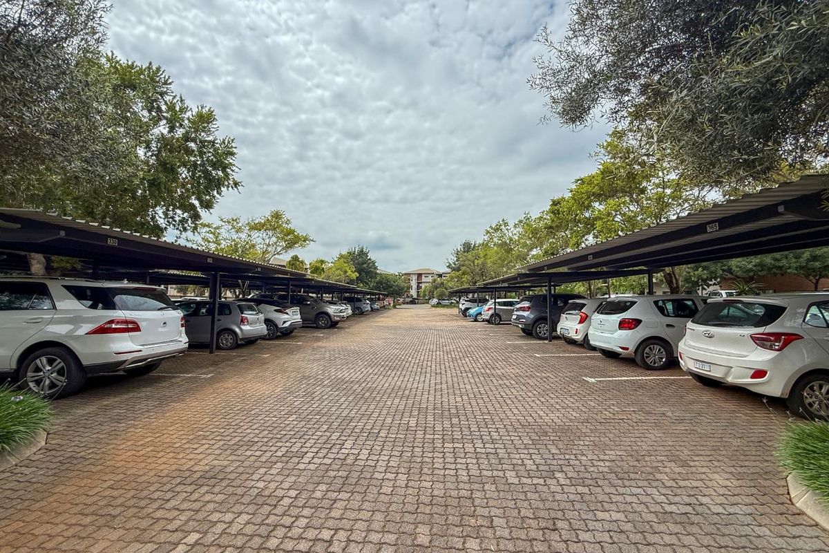 A paved outdoor parking area featuring covered parking bays with metal roofing structures, surrounded by mature trees.