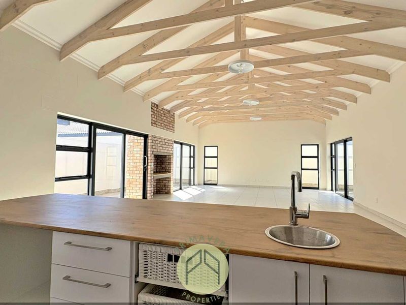 Spacious open-plan living area featuring exposed timber trusses, a built-in brick fireplace, and a modern kitchen island with a sink.
