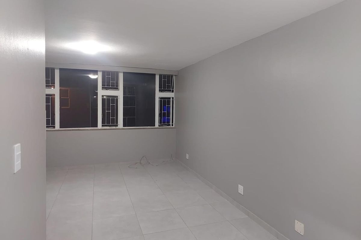 A spacious, empty living area featuring neutral grey walls, tiled flooring, and large windows with security bars.