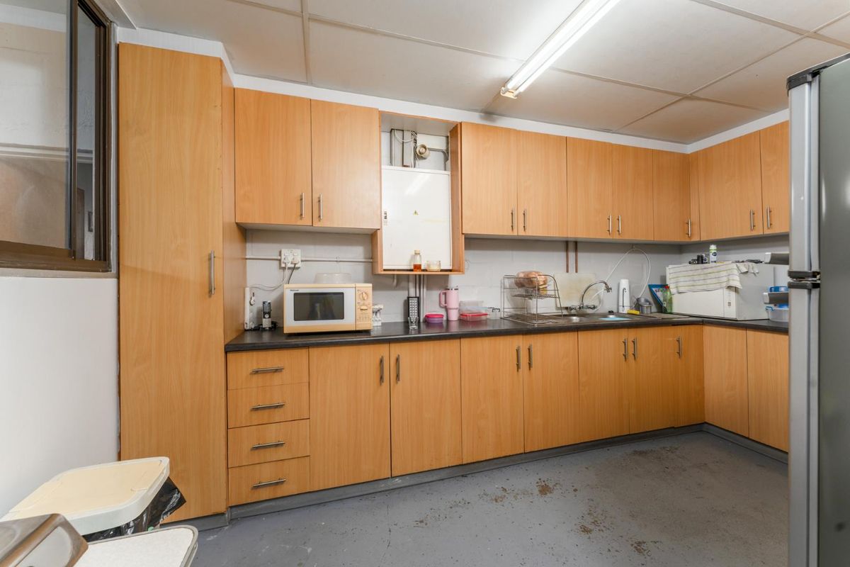 A functional office kitchenette featuring light wood-tone cabinetry, laminate countertops, a stainless steel sink, and overhead fluorescent lighting.