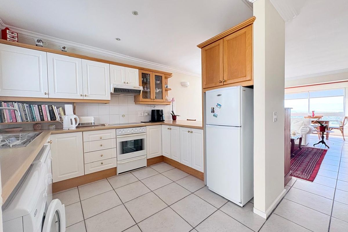 A functional kitchen featuring white and timber cabinetry, integrated oven and hob, and tiled flooring, flowing into an adjacent living area with ocean views.