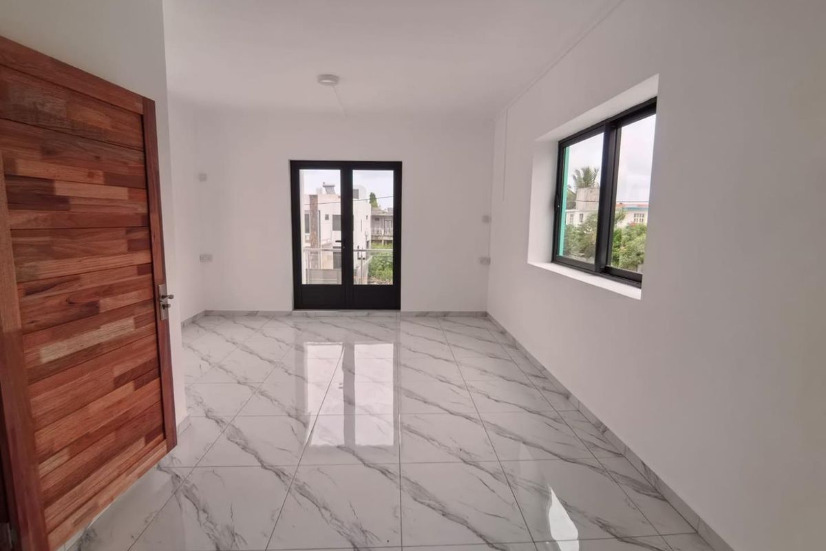 A bright, empty living room featuring polished marble-look floor tiles, a solid wood door, and black-framed windows leading to an exterior balcony.