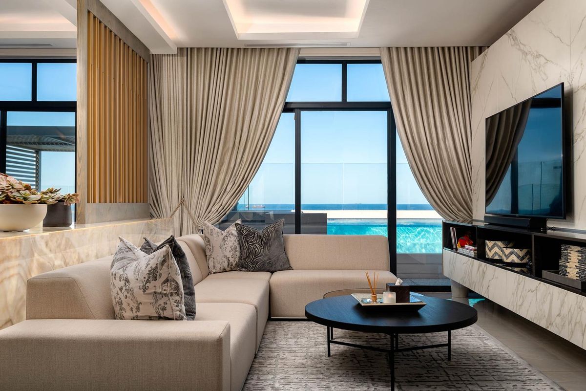Modern living room featuring a marble-clad media wall, recessed ceiling lighting, and floor-to-ceiling glass doors opening to a private balcony with pool and sea views.