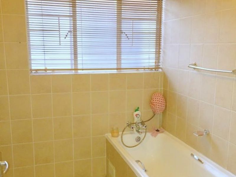A dated bathroom featuring a built-in bathtub with wall-mounted mixer tap, tiled surfaces, and a window with fitted blinds.