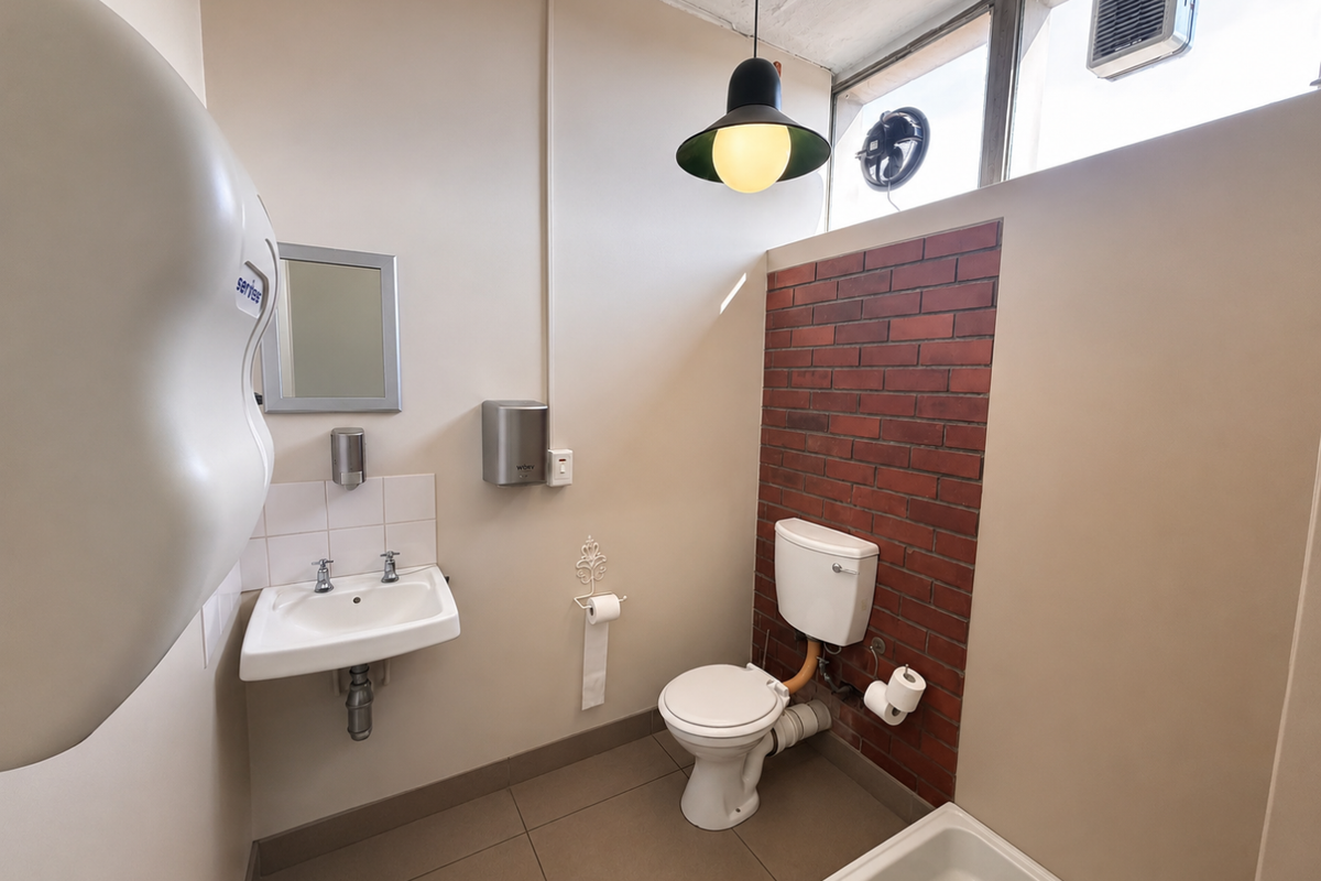 Functional industrial-style restroom featuring a wall-mounted basin, toilet with exposed brick accent wall, and high-level ventilation window.