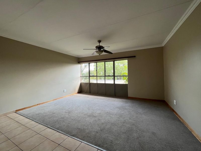 A spacious, empty living room featuring neutral wall tones, a ceiling fan, and a combination of carpet and tile flooring with large windows.