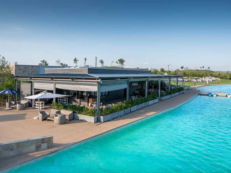 A modern clubhouse facility featuring a covered patio area overlooking a large, clear swimming pool with paved decking and landscaped surroundings.