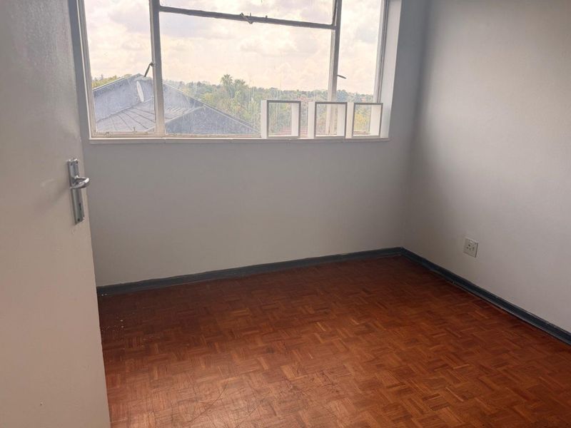 An empty bedroom featuring original parquet flooring, a large metal-framed window, and neutral wall finishes, offering a blank canvas for renovation.