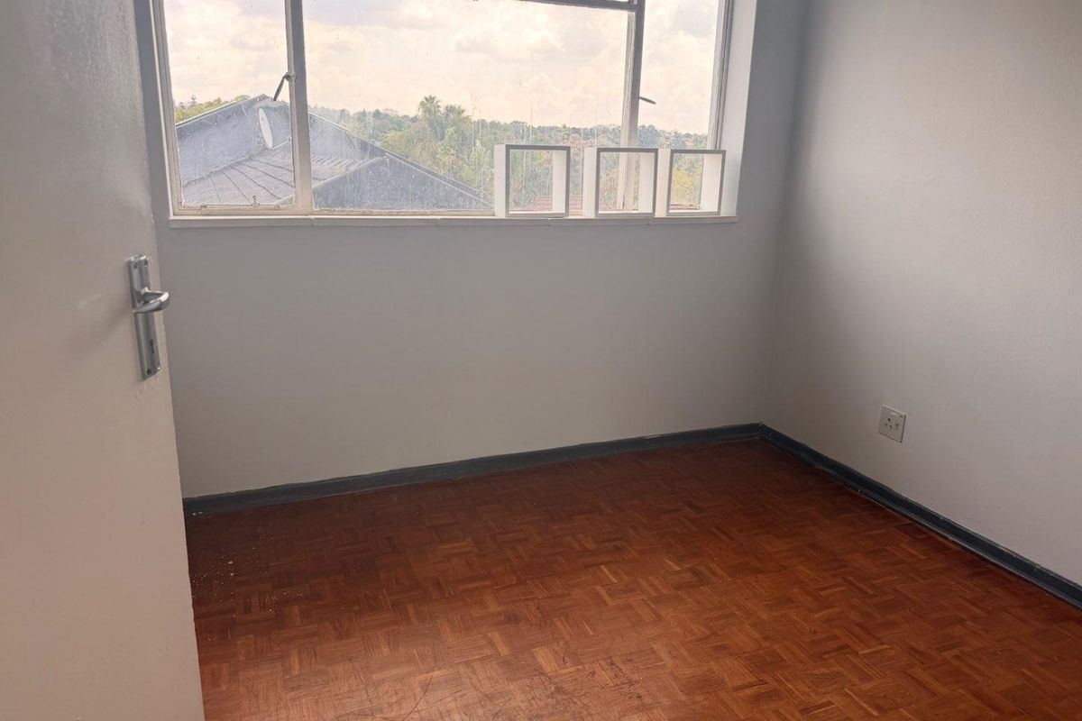 An empty bedroom featuring original parquet flooring, a large metal-framed window, and neutral wall finishes, offering a blank canvas for renovation.