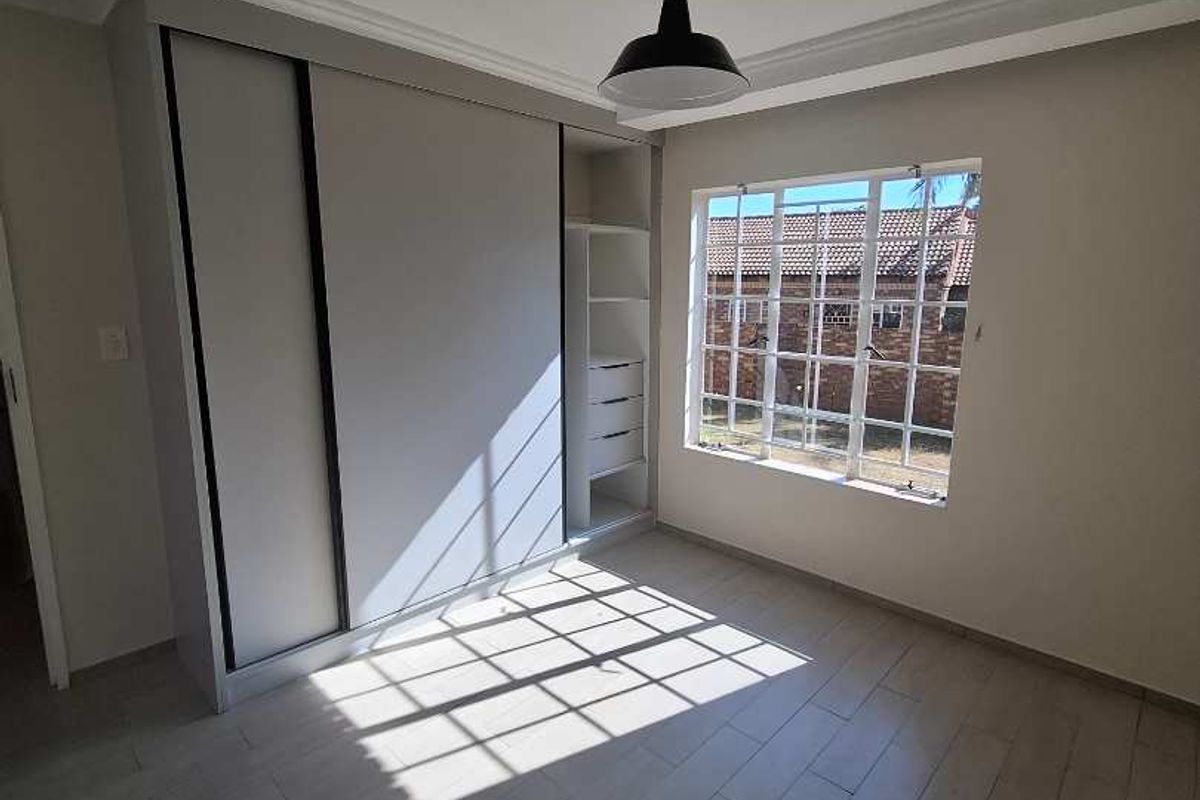 A bright bedroom featuring modern light-toned wood-look flooring, a built-in sliding door wardrobe with shelving, and a large window providing natural light.