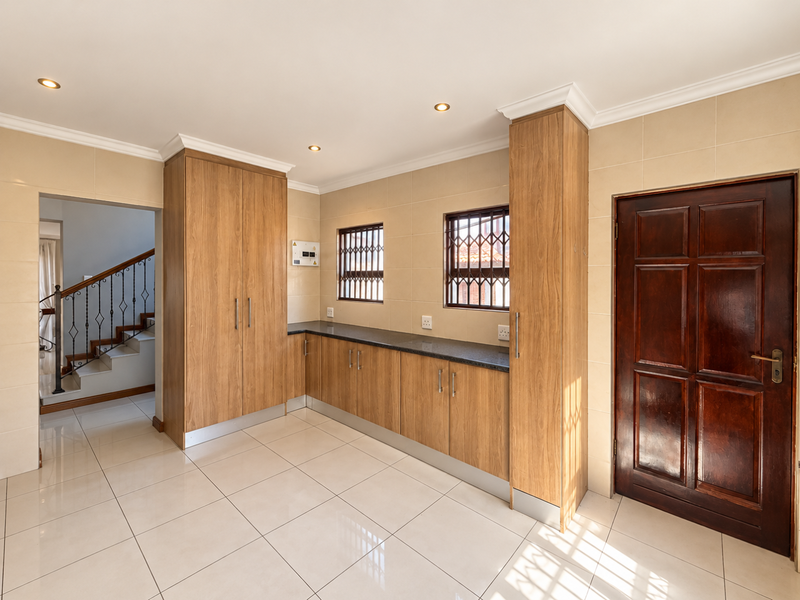 A clean and functional scullery area featuring built-in wood-finish cabinetry, granite countertops, tiled flooring, and direct access to the main living area and exterior.