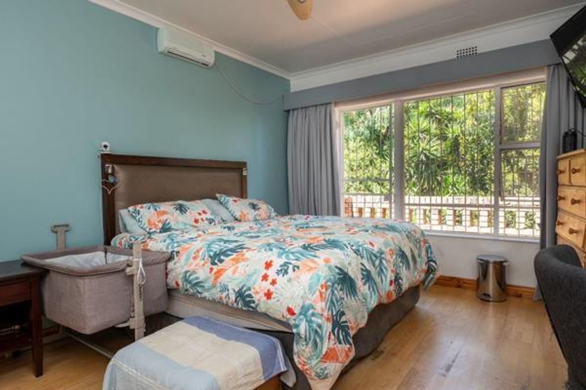 A spacious bedroom featuring light wood flooring, a wall-mounted air conditioning unit, and large windows with security bars overlooking the garden.