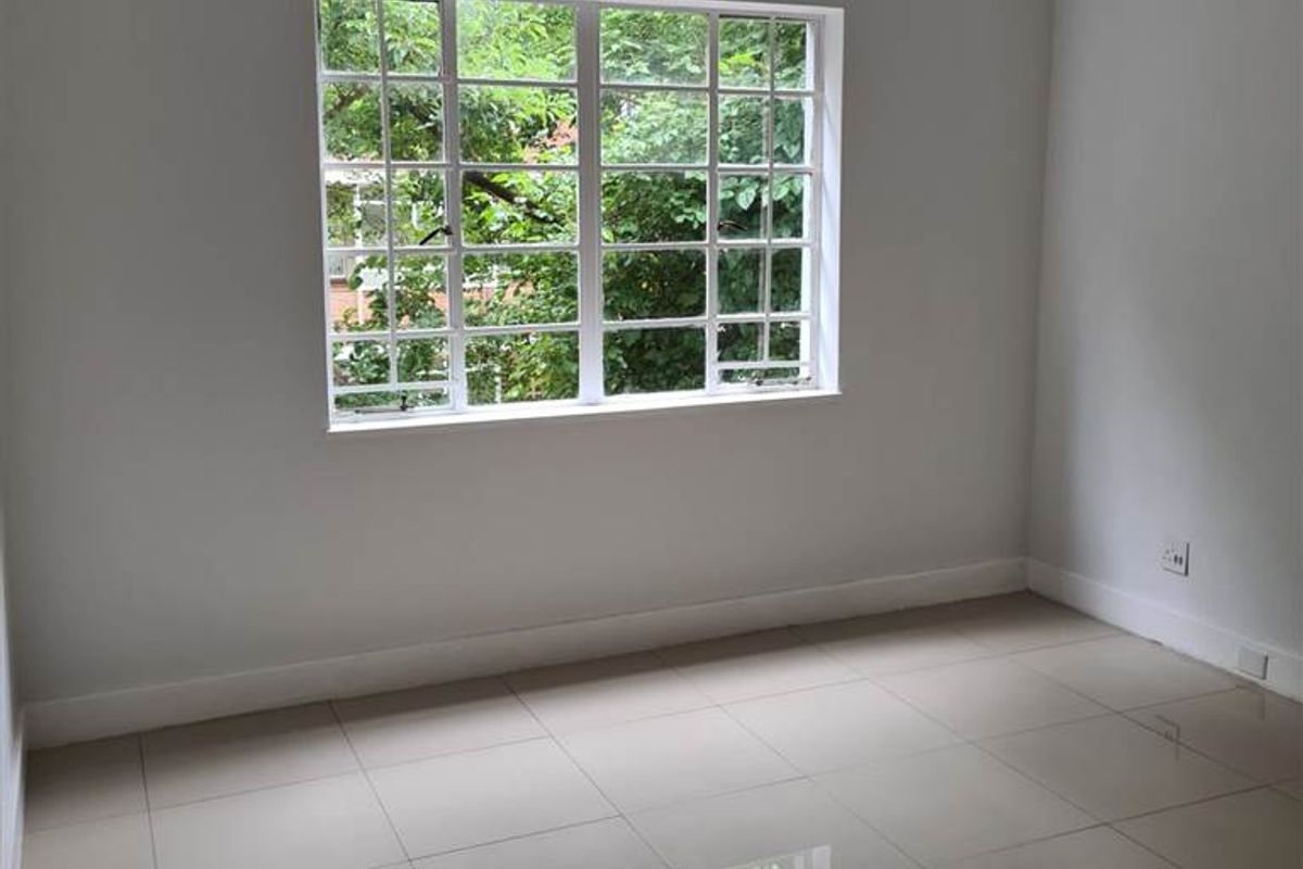 A bright, empty bedroom featuring neutral tiled flooring and a large multi-pane window overlooking greenery.