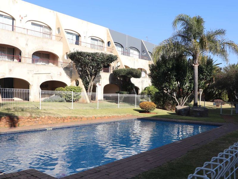 Communal swimming pool area featuring brick paving and metal fencing, set against the backdrop of a multi-story apartment building with private balconies.