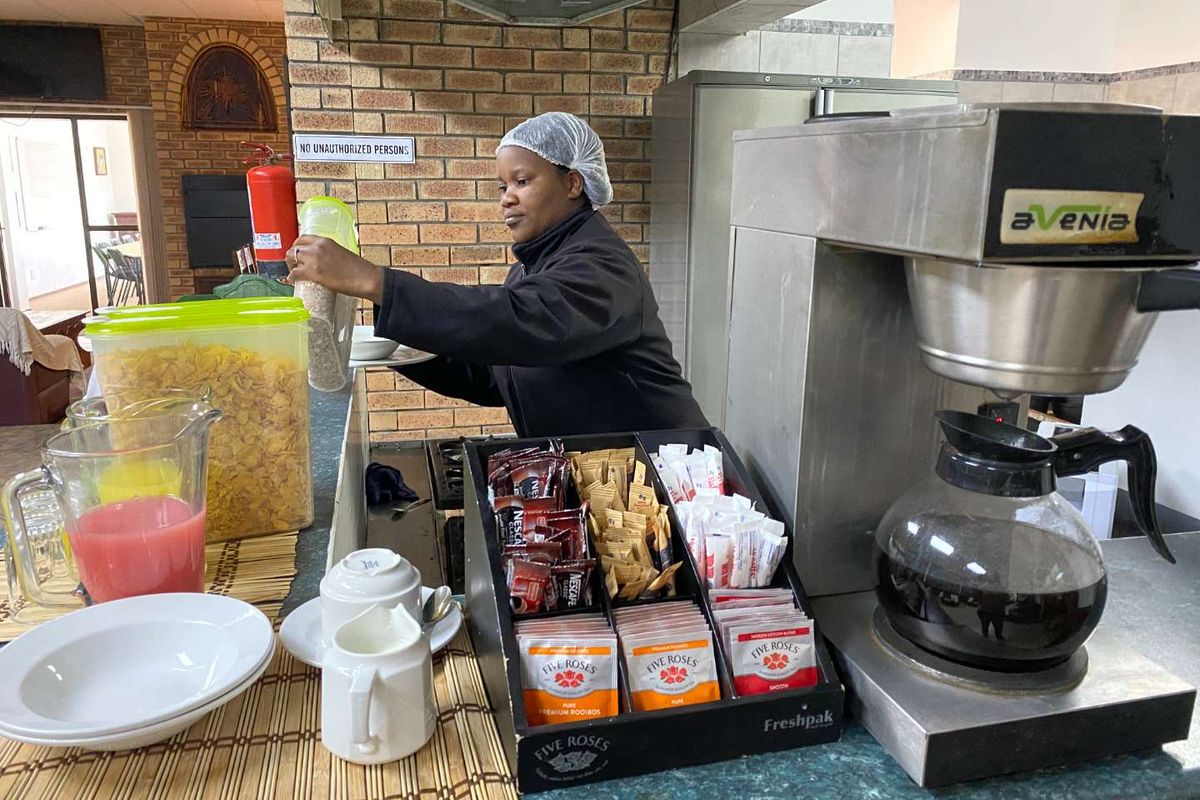 A functional breakfast service area featuring a brick-accented wall, laminate countertop, and integrated coffee brewing station within the guest house kitchen.