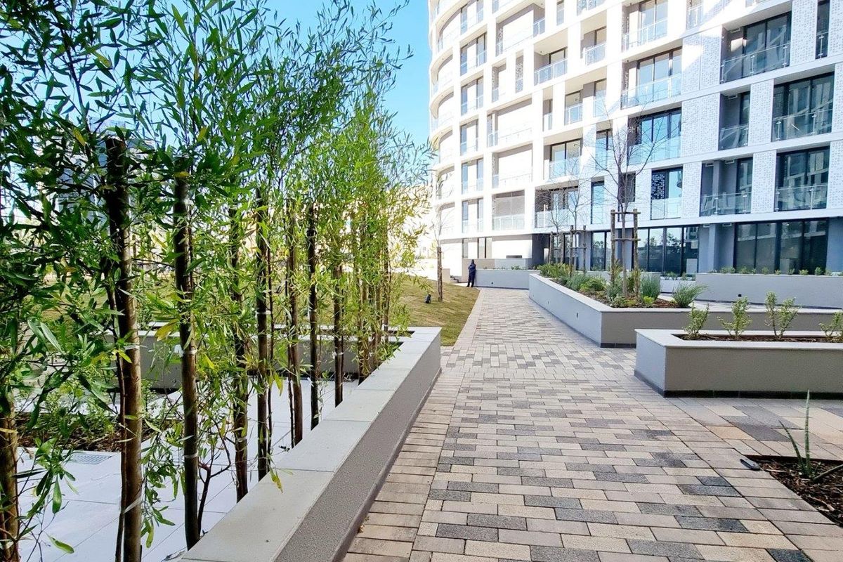 A well-maintained outdoor communal area featuring paved walkways, modern raised concrete planters, and landscaped greenery adjacent to a contemporary apartment building.