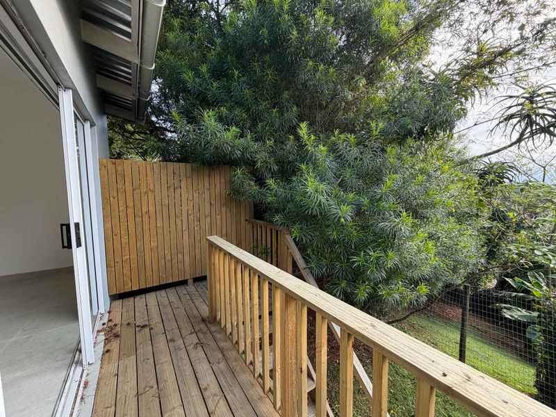 A wooden balcony featuring a timber balustrade and privacy screen, accessible via sliding glass doors, overlooking a lush garden area.