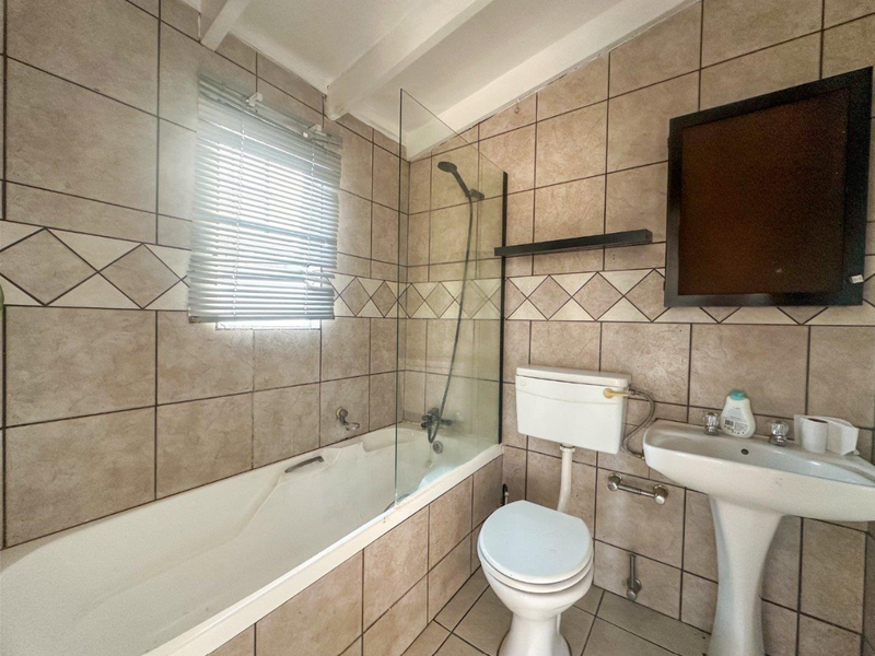 A functional bathroom featuring a bathtub with a glass shower screen, pedestal basin, and toilet, finished with neutral wall and floor tiling.