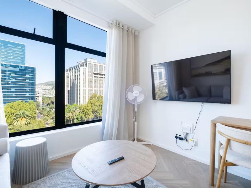 Bright living area featuring large floor-to-ceiling windows with city views, herringbone wood flooring, and a wall-mounted television setup.