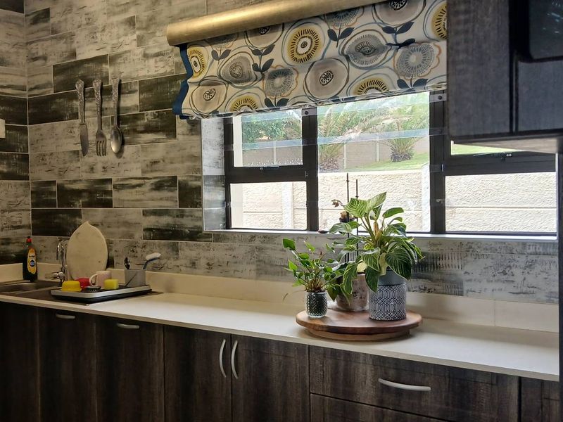 A functional kitchen area featuring dark wood-look cabinetry, a light-toned countertop, and a distinctive textured tiled splashback with a window view.