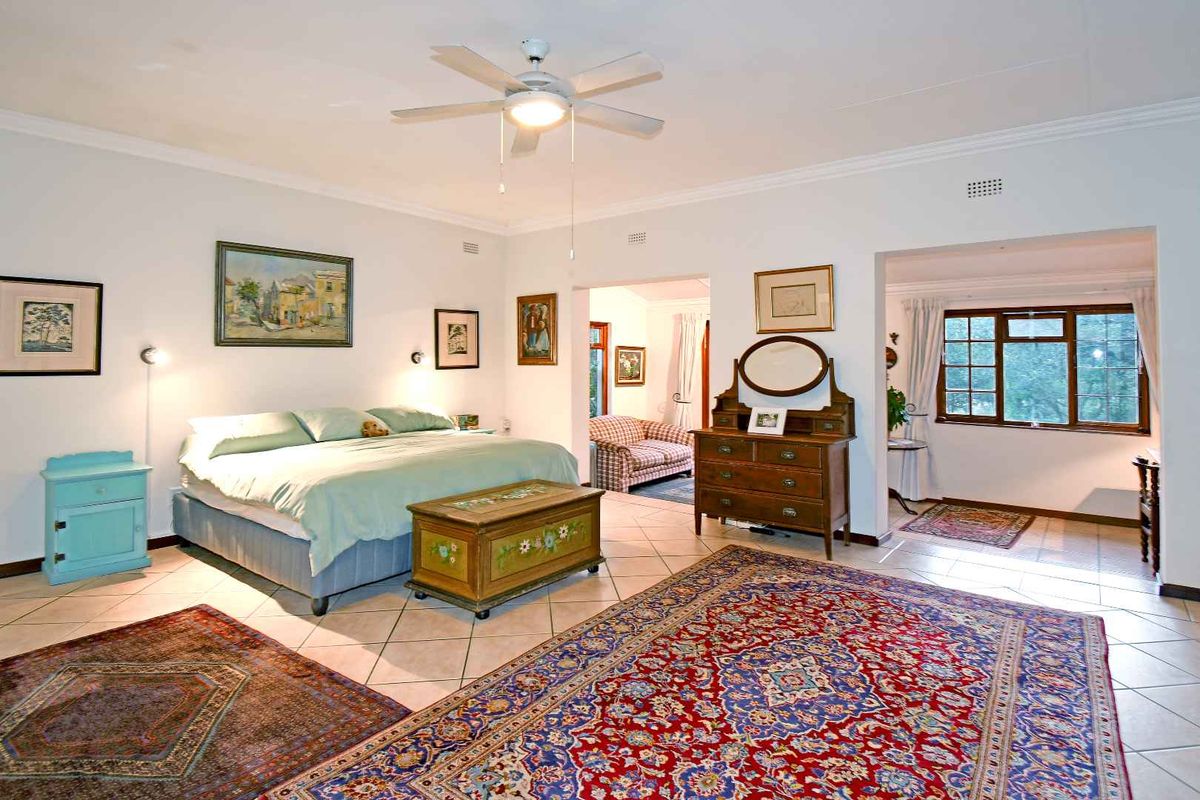 Spacious bedroom featuring tiled flooring, a ceiling fan, and an open-plan layout connecting to an adjacent sitting area with large windows.