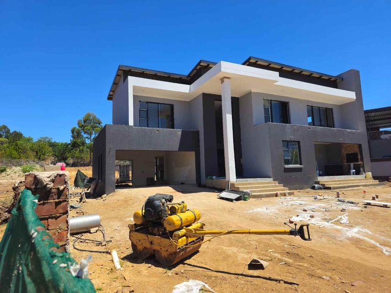 A modern double-storey house under construction, featuring a contemporary facade with grey and white finishes, covered entrance, and integrated garage space.