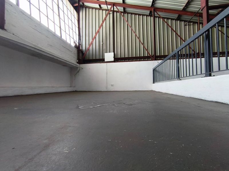 An empty industrial mezzanine level featuring exposed steel trusses, corrugated metal cladding, large steel-framed windows, and a protective metal railing.